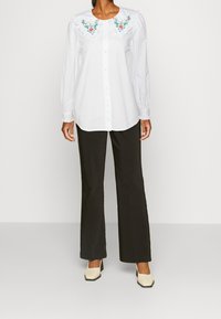 Camicia bianca con colletto ricamato di grandi dimensioni con motivi floreali, abbinata a pantaloni neri a zampa d'elefante e scarpe con tacco a blocco di colore bianco sporco.