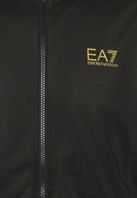 Giacca nera con zip in tessuto liscio. Presenta un logo EA7 Emporio Armani dorato di grande rilievo e una chiusura con zip a contrasto.