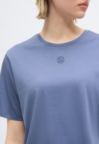 Γυναίκα που φοράει ανοιχτό μπλε T-shirt με στρογγυλή λογότυπο στο κέντρο του στήθους, κοντοκομμένο κάτω από το σαγόνι.