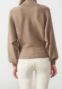 Pull en laine marron à côtes avec un col montant, des manches ballons et un ourlet ajusté, associé à un pantalon blanc ajusté. Texture lisse avec un design subtil.