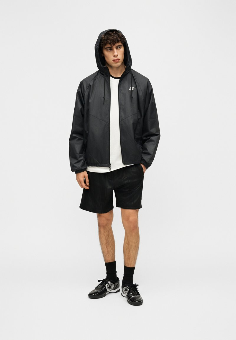 Giovane uomo con giacca nera con cappuccio, maglietta bianca, pantaloncini neri, calzini neri e scarpe da ginnastica nere e bianche in piedi davanti a uno sfondo semplice.