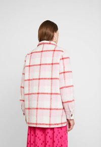 Tarkoitettu karvatuotteille, valkoisen ja vaaleanpunaisen sävyissä. Koristeena punainen ruutukuosi ja napit hihoissa. Näyttää takapuolelta.