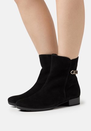 Botines de ante negro con una textura suave y tacón plano. Cuentan con una cadena decorativa dorada en el lado exterior. Diseño limpio, sin adornos.