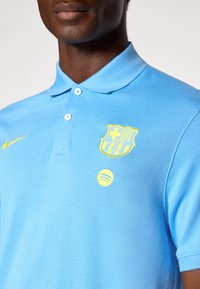 Svetlo modra polo majica z mehko teksturo, ki ima rumen logotip FC Barcelone, rumeni Nike swoosh in rumen logotip Spotify.