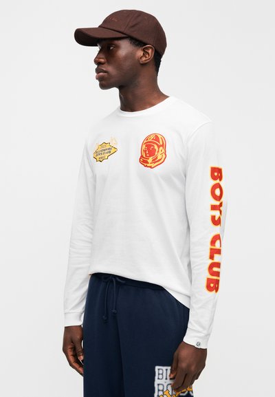 Λευκό πουκάμισο με μακριά μανίκια που διαθέτει κίτρινες και κόκκινες εκτυπώσεις αστροναύτη και τη φράση «Boys Club»· φορεμένο με σκούρο μπλε παντελόνι. Καφέ καπέλο συμπληρώνει το σύνολο.