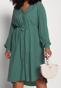 Tyrkysové šaty na knoflíky s výstřihem do V, dlouhými rukávy, páskem v oblasti pasu a kapesní. Doplněno světlou béžovou strukturovanou kabelkou s kruhovou rukojetí.