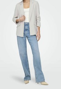 Blazer gris clair à épaules structurées, devant ouvert et manches retroussées, associé à un jean taille haute bleu clair et des chaussures pointues beiges.