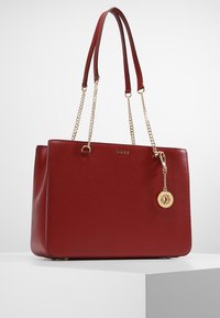 Punainen rakenteellinen olkalaukku, joka on valmistettu teksturoidusta nahasta, kahdella olkahihnalla, kullanvärisillä ketjutyylisillä yksityiskohdilla ja pyöreällä logoriipuksella.