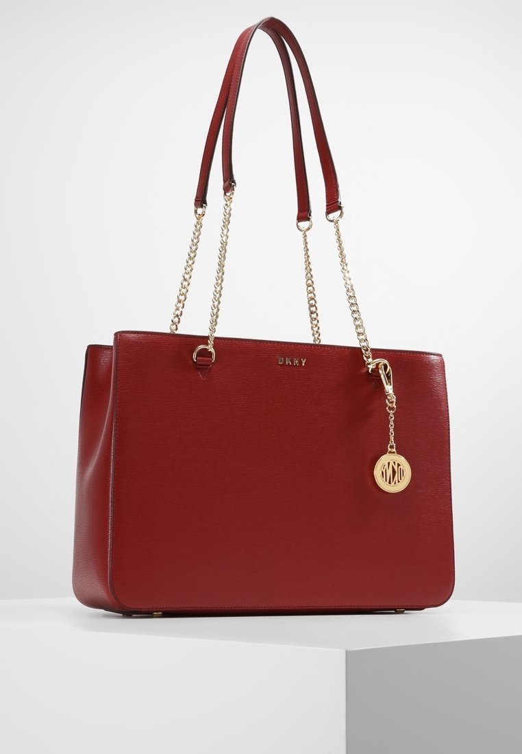 Punainen rakenteellinen olkalaukku, joka on valmistettu teksturoidusta nahasta, kahdella olkahihnalla, kullanvärisillä ketjutyylisillä yksityiskohdilla ja pyöreällä logoriipuksella.