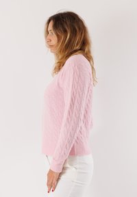 Una donna con capelli ondulati, in un maglione di lana rosa e pantaloni bianchi, è in piedi di profilo verso sinistra su uno sfondo chiaro e semplice.