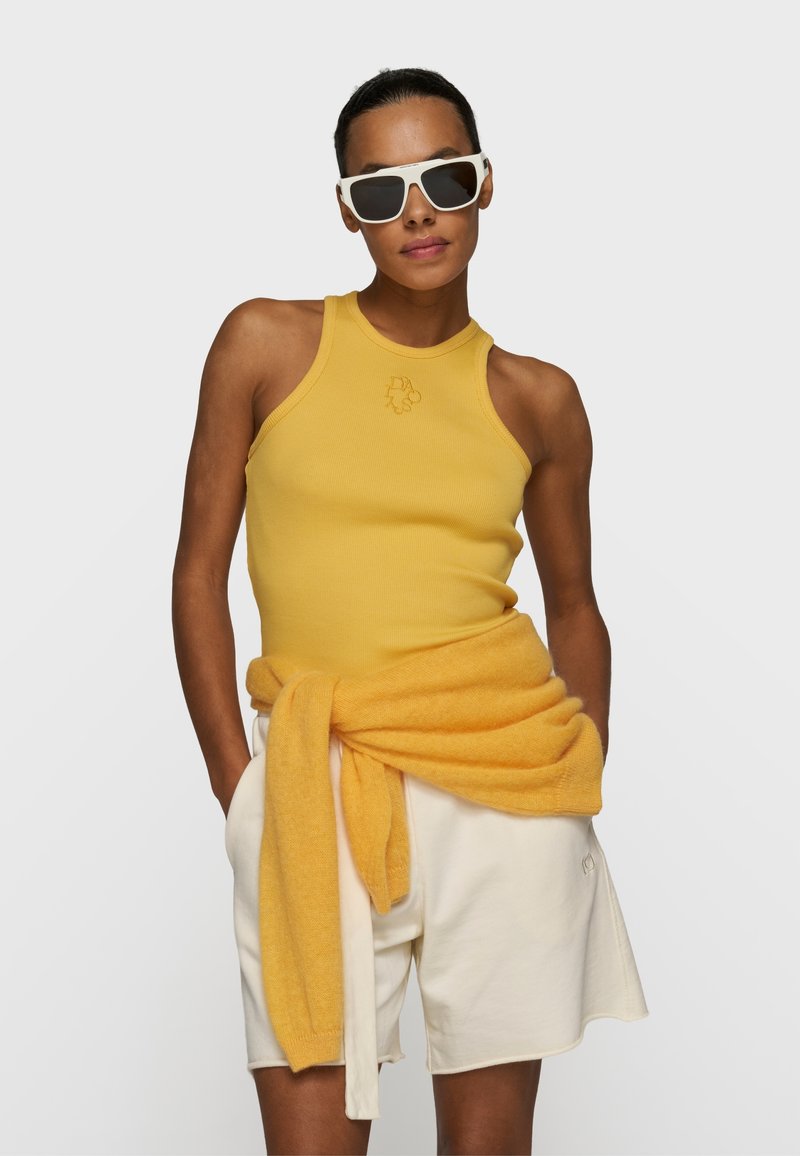 Femme portant des lunettes de soleil blanches, un haut sans manches jaune et un short blanc avec un pull jaune noué autour de la taille, sur un fond uni.