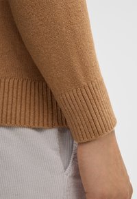 Brązowy sweter z dzianiny z rękawem zakończonym ściągaczem. Miękka tekstura z delikatnym wzorem dzianiny, w połączeniu z jasnoszarymi spodniami z korduroy widocznymi poniżej.