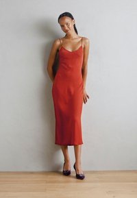 Abito in satin rosso con spalline sottili, lunghezza a metà polpaccio, abbinato a tacchi viola con decorazioni, in piedi su un pavimento di legno contro un muro semplice.