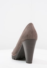 Zapato de tacón alto de ante marrón con un diseño afilado, tacón texturizado y punta redondeada. Se sitúa sobre una suela sólida y con patrón para mayor tracción.
