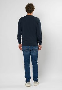 Marineblauwe lange mouwen trui met ribbelmanchetten en tailleband, gecombineerd met blauwe jeans en lichtgekleurde sneakers. Model van achteren weergegeven.