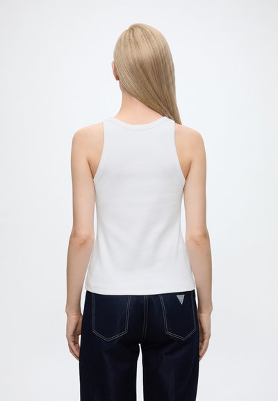 Personne blonde portant un haut blanc sans manches et un jean bleu foncé vue de dos sur un fond uni.