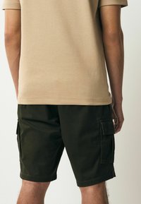 Beżowy t-shirt z krótkim rękawem w zestawieniu z ciemnozielonymi szortami cargo posiadającymi kieszenie boczne i prosty krój. Gładka tekstura materiału.