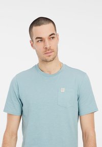 Svetlomodré tričko s krátkymi rukávmi vyrobené z mäkkej látky, s malou náprsní vreckom a štítkom, okrúhlym výstrihom a voľným strihom.