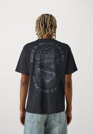 Personne aux cheveux blonds torsadés portant un t-shirt noir avec un graphisme du Cœur immaculé de Marie et un texte religieux environnant.