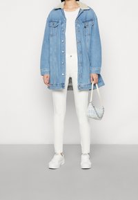Persona que lleva una chaqueta de denim azul claro con un cuello de borreguito blanco, pantalones blancos, zapatillas blancas y sosteniendo un pequeño bolso de cuadros azules.