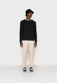 Suéter negro con dobladillo acanalado, camiseta crema debajo, pantalones beige y gorra de cubo negra. Un par de zapatillas negras y blancas.