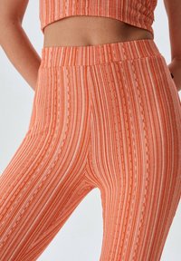 Oranje elastische legging met dunne witte verticale strepen en een gestructureerd ribbelpatroon. Beschikt over een hoge tailleband en een aansluitend ontwerp.