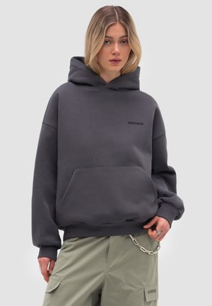 Frau mit blonden Haaren, die einen dunkelgrauen, übergroßen Hoodie und hellgrüne Cargo-Hosen mit einer Kettenverzierung vor einem einfarbigen Hintergrund trägt.