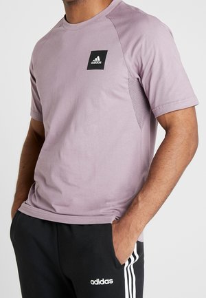 Homme portant un t-shirt Adidas violet clair avec un logo noir, un pantalon Adidas noir avec des bandes blanches et un logo, les mains dans les poches.