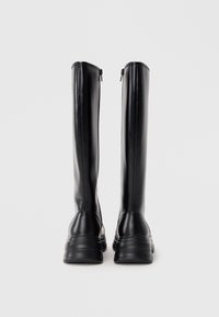 Ζεύγος ψηλών μαύρων δερμάτινων μπότες με χοντρούς πάτους, εμφανιζόμενες από την πίσω πλευρά σε απλό ανοιχτό γκρίζο φόντο.