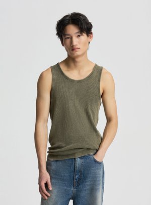 Młody mężczyzna z czarnymi włosami, ubrany w oliwkowo-zielony tank top i niebieskie dżinsy, stoi z jedną ręką w kieszeni na jednolitym tle.