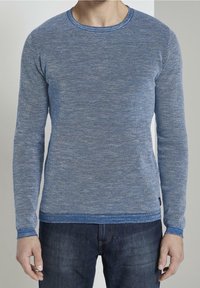 Maglione lavorato a maglia blu con maniche lunghe, design testurizzato, scollo a coste e polsini. Abbinato a jeans di denim scuro.