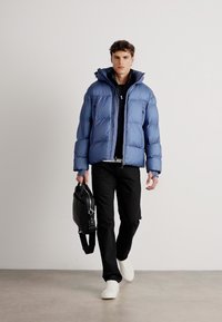 Giacca a vento blu con finitura lucida, abbinata a un maglione nero e pantaloni. Il modello porta una borsa nera e indossa scarpe da ginnastica bianche.