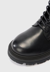 Musta nahkasaapas paksulla kumipohjalla ja mustilla nauhoilla vaaleaa taustaa vasten, esittäen etu- ja sivupintoja.