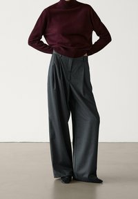 Person in burgunderfarbenem Rollkragenpullover und hochgeschnittenen, dunkelgrauen, gestreiften, weiten Hosen mit schwarzen, spitzen Schuhen.