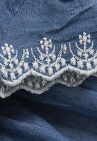 Denimowy materiał z białym haftem wzdłuż dołu, z kwiatowymi wzorami i falbaniastymi brzegami, ukazujący teksturowaną powierzchnię.