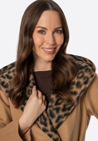 Beżowy płaszcz z szalowym kołnierzem w panterkę, wykonany z miękkiego materiału. Modelka prezentuje płaszcz z brązową bluzką, podkreślając teksturę i wzór.