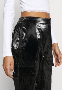Black, shiny, synthetic cargo pants with a gathered waistband and patch pockets; paired with a white long-sleeve top featuring lace trim.