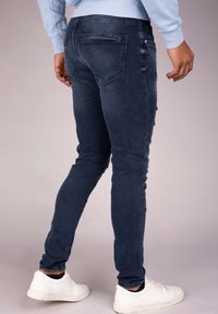 Slim-fit denim jeans in dark blue with a slight faded effect, featuring two back pockets and a distressed detail on the side. Paired with white sneakers.
