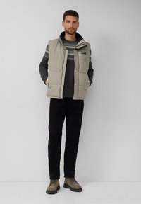 Homme debout les mains dans les poches, portant un gilet matelassé beige, un pull à motifs, un pantalon noir et des bottes à lacets marron sur fond uni.