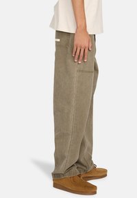 Olivgrüne, weit geschnittene Hose aus weichem, strukturiertem Stoff mit Seitentaschen und einem Etikett auf der Rückseite. Kombiniert mit braunen Slippern.