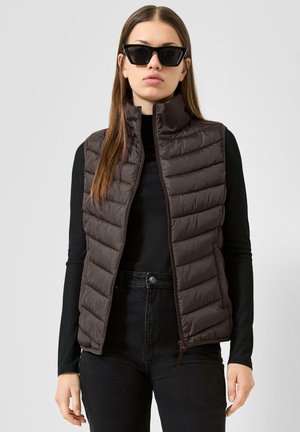 Frau trägt schwarzes Langarmshirt, dunkelbraune gesteppte Weste, schwarze High Waist Jeans und schwarze Sonnenbrille mit Katzenaugenform, steht vor weißem Hintergrund.