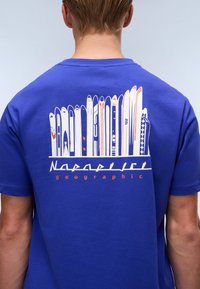 Blå kortærmet t-shirt med et hvidt og orange grafisk design af skitegn på ryggen, med "Napapijri geographic" tekst nedenunder.