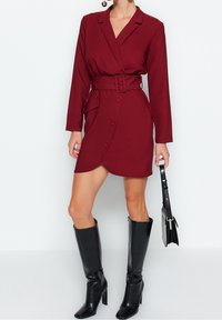 Burgundy omlottklänning med långa ärmar, bälte vid midjan, framknappar och asymmetrisk nederkant. Kombineras med svarta knähöga stövlar och en handväska.