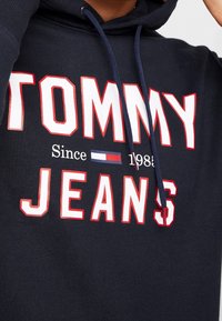 Námořnická mikina s textem "TOMMY JEANS Since 1985" a červeno-bílo-modrým logem vlajky Tommy Hilfiger uprostřed hrudi.