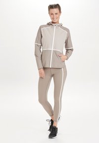 Lätt beige jacka med vita reflexband, dragkedja framtill, huva och sidofickor, ihop med matchande leggings och svarta sportskor.