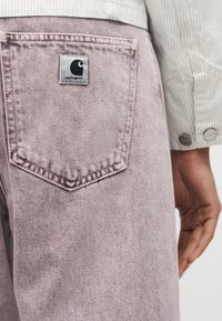 Πίσω τσέπη ξεθωριασμένου μωβ τζιν με κεντημένη ετικέτα λογότυπου Carhartt, δίπλα σε ένα χέρι και μανίκι μπεζ κορδουρένιου μπουφάν με μεταλλικά κουμπιά.