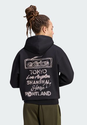 Person med dreadlocks bundet op bærer en sort Adidas-hoodie med bynavnene Tokyo, Los Angeles, Shanghai, Herzo og Portland trykt på bagsiden.