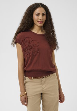Kvinde med langt krøllet hår iført en brun kortærmet top med et stort blomsterdesign og beige bukser med et flettet bælte.