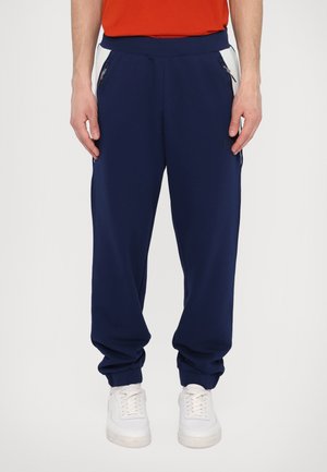 Mørkeblå joggebukser med elastisk midje og mansjetter, med sidelommer med glidelås og kontrasterende hvite sidestykker. Glatt tekstur.