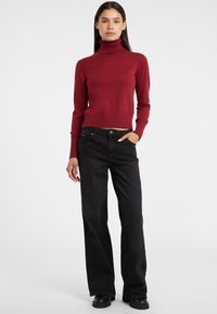 Maglione rosso a collo alto, corto, con orlo a costine abbinato a jeans larghi neri e stivali chunky neri. Design semplice, texture liscia.